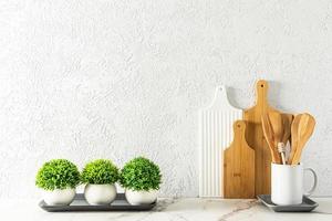 cutting boards and wooden spoons in a white mug on a marble white countertop against a gray cement wall with green houseplants. photo