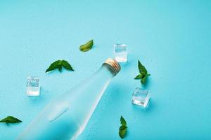 una botella de agua helada, cubitos de hielo, gotas y hojas de menta sobre un fondo azul. foto