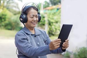 Happy Asian senior woman wears headphones, holds smart tablet, listen to music. Concept, elderly use technology smart device. Relax with online music, watch vedio. Learning new thing from internet. photo