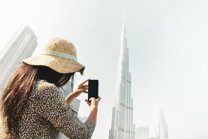 joven turista caucásica toma una foto móvil de burj khalifa con un teléfono inteligente en un día soleado y nublado