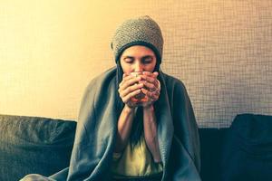 una mujer caucásica preocupada y triste se sienta bajo una manta en un sofá con una taza de té, aumentando los costos en los hogares privados por la factura del gas debido a la inflación y la guerra, la crisis energética foto