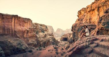 una joven turista caucásica se sienta y sonríe mientras mira las hermosas tallas de la ciudad antigua de petra. Visita famosa destino de atracción de Jordania. panorama del sitio arqueológico de petra