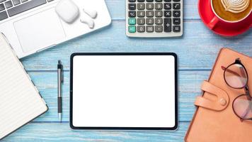 tableta con pantalla en blanco, computadora portátil, calculadora, cuaderno, anteojos, bolígrafo y taza de café en el escritorio de madera, vista superior plana. foto