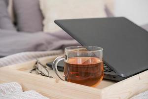 portátil y taza de té en una bandeja en la cama del dormitorio. trabajo desde casa, aprendizaje a distancia o un pasatiempo cómodo en línea. foto