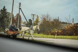 Pabrade, Lithuania, 2022 - View from a car on an old horse-driven carriage with couple in it crossing a small town road photo
