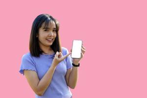 mujer asiática sonriente jugando y actualizando nuevas aplicaciones. su mano apuntó al teléfono con el área de trabajo de la pantalla en blanco charlando sobre fondo rosa. enfoque suave y selectivo.