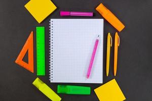 Back to school. On black table lies notebook checkered with pencil for text with empty space. All around are green, yellow, pink, orange stationery. Rulers, markers, felt tip pens, pens, adhesive note photo