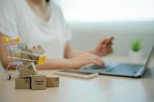 Online shopping-boxes or parcels are placed on the table and shopping carts. Blurred background, woman use credit card to make online purchases on laptop. Online service. New normal shopping delivery. photo