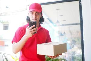 el repartidor con uniforme rojo ve la información del cliente en el smartphone con la caja del paquete en la mano. servicio de entrega rápida foto