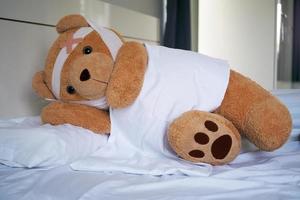 Teddy bear lying sick in bed With a headband and a cloth covered photo