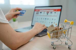 Online shopping-boxes or parcels are placed on the table and shopping carts. Blurred background, woman use credit card to make online purchases on laptop. Online service. New normal shopping delivery. photo