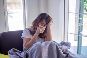 mujer de pelo largo sentada en el sofá sufre de gripe, tos y estornudos. sentarse en una cobija debido a la fiebre alta y taparse la nariz con un pañuelo de papel porque estornuda todo el tiempo. foto