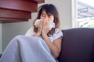 Asian woman sat ill on the sofa in the house, having fever, coughing, sneezing, and stuffy nose. Covering the nose with a piece of paper. photo