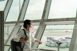 un hombre asiático sostiene una mochila en la ventana del aeropuerto mientras espera un vuelo en la puerta de embarque antes de la salida. foto