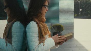 una joven morena seria con anteojos se encuentra frente a una pared de espejo azul con libros en las manos en la calle video