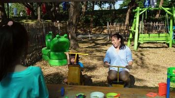 des sœurs heureuses jouent avec de la nourriture et des jouets d'épicerie en bois sur l'aire de jeux extérieure avec sa mère. jolie fille asiatique jouant un rôle dans la vente de jus de fruits au parc. la famille passe du temps ensemble en vacances. video