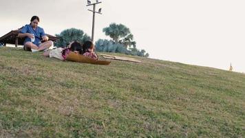 Lycklig systrar spelar på de parkera diabilder ner från de gräs- kulle Sammanträde på en kartong låda. Lycklig barn spelar utomhus i sommar. familj utgifterna tid tillsammans på semester. video