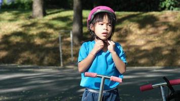 retrato de una joven activa con casco preparándose para andar en moto por la carretera en un parque al aire libre el día de verano. niña asiática feliz montando un scooter a la escuela. video