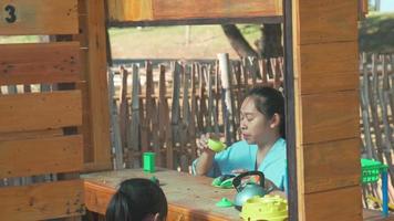 irmãs felizes brincam com comida e brinquedos de mercearia de madeira no playground ao ar livre com a mãe. linda garota asiática interpretando vendendo suco de frutas no parque. família passando tempo juntos nas férias. video