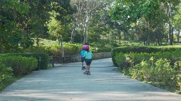 retrato de una niña activa montando scooter en la calle en un parque al aire libre el día de verano. feliz chica asiática con casco montando una patineta en el parque. ocio activo y deporte al aire libre para niños. video