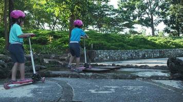 portrait d'une petite fille active faisant du scooter dans la rue dans un parc extérieur le jour d'été. heureuse fille asiatique portant un casque faisant du scooter dans le parc. loisirs actifs et sports de plein air pour enfant. video