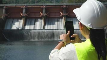 Female engineer in white hat working with smartphone and looking away at dam with hydroelectric power plant and irrigation. video