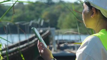 miljö- ingenjör bär en vit hjälm användningar en mobil telefon till spela in data analyserar syre nivåer i en reservoar. vatten och ekologi begrepp. video