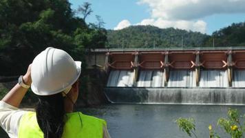 Female engineer wearing green vest and white helmet working outside dam with hydroelectric power plant and irrigation. Renewable energy system. Sustainable energy concept.