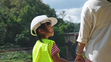 ingenjör mor innehav henne dotters hand och leende på varje Övrig mot de bakgrund av en damm med en vattenkraft kraft station. begrepp av förnybar energi och kärlek av natur och familj. video