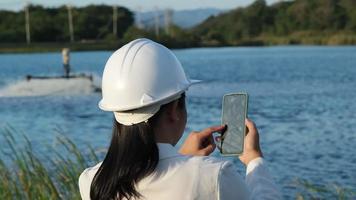 miljö- ingenjör bär en vit hjälm användningar en mobil telefon till spela in data analyserar syre nivåer i en reservoar. vatten och ekologi begrepp. video