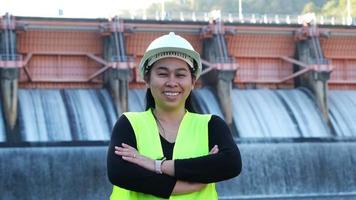 sorridente femmina ingegnere nel verde veste e casco in piedi al di fuori contro sfondo di idroelettrico diga, paratoia con fluente acqua attraverso cancello. video