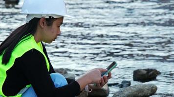 Female environmentalist using mobile phone to record water analysis data in dam. Researchers are collecting water samples from the river to test and detect pathogens. Water and ecology concept