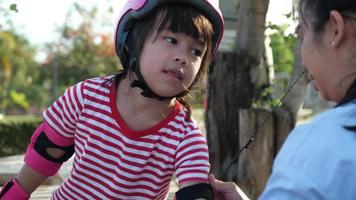 la joven madre ayuda a su hija a ponerse las almohadillas protectoras y el casco antes de patinar en el parque. ocio activo y deporte al aire libre para niños. video