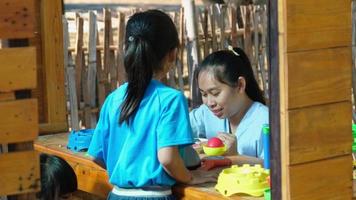 Happy sisters play with food and wooden grocery toys at the outdoor playground with her mother. Cute Asian girl roleplaying selling fruit juice at the park. Family spending time together on vacation.