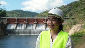 Smiling female engineer in green vest and helmet standing outside against background of hydroelectric dam, floodgate with flowing water through gate.