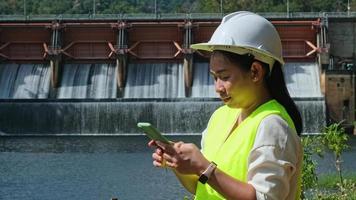 engenheira de chapéu branco trabalhando com smartphone e desviando o olhar para represa com usina hidrelétrica e irrigação. video