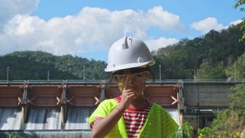 Portrait of a little girl engineer wearing a green vest and white helmet smiling happily on the background of the dam. video