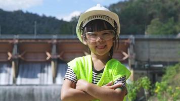 Porträt eines kleinen Ingenieurmädchens, das eine grüne Weste und einen weißen Helm trägt und glücklich auf dem Hintergrund des Damms lächelt. video