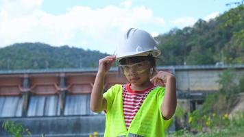 porträtt av en liten flicka ingenjör bär en grön väst och vit hjälm leende lyckligt på de bakgrund av de damm. video