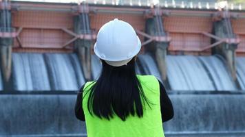 Rear view of female engineer in green vest and helmet standing outside against background of dam with hydroelectric power plant and irrigation. video