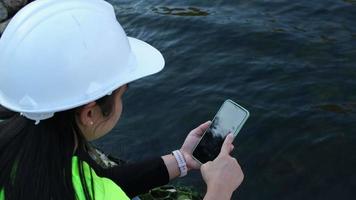 femmina ambientalista utilizzando mobile Telefono per disco acqua analisi dati nel diga. ricercatori siamo raccolta acqua campioni a partire dal il fiume per test e individuare agenti patogeni. acqua e ecologia concetto video