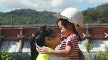 Engineer mother, wearing helmet to her daughter against background of dam with hydroelectric power plant. Concepts of environmental engineering, renewable energy and love of nature and family.