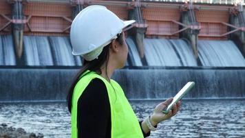 Female environmentalist wearing green vest and safety helmet working outdoors against the background of hydroelectric dam and recording analytical data for water contamination on mobile phone. video