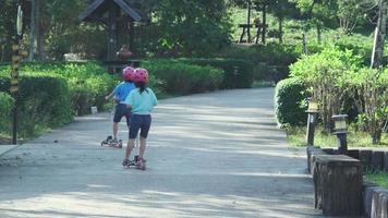 porträtt av aktiva liten flicka ridning skoter på gata i utomhus- parkera på sommar dag. Lycklig asiatisk flicka bär en hjälm ridning en sparka skoter i de parkera. aktiva fritid och utomhus- sport för barn. video