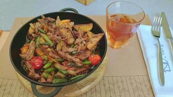 déjeuner de chevreuil chaud dans une poêle à frire. morceaux de viande juteux avec des légumes sur la table en gros plan et un verre de jus. plat contient des pommes de terre cuites, des oignons, des poivrons, des herbes et des asperges dans de l'huile bouillante. video