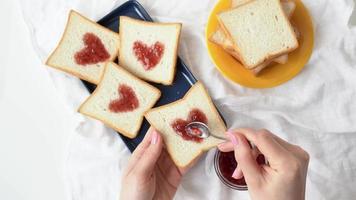 Scheibe Brot mit Erdbeermarmelade in Herzform in der Mädchenhand. frau, die ein süßes frühstück kocht - leckere sandwiches zum valentinstag. Frauenhände tragen herzförmige rote Konfitüre auf weißen Toast auf video