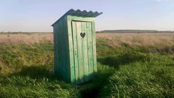 Alte Holztoilette mit einem geschnitzten Fenster mit der Form eines Herzens, das an der Tür ausgeschnitten ist, auf freiem Feld. Vintage-WC. Eine rustikale grüne Toilette im Freien in einer Feldlandschaft aus Gras im Wind bei Sonnenaufgang.