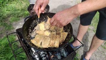 rostning gädda i en panorera på brand. små Krispig bitar av fisk är friterad i olja. de begrepp av matlagning mat i natur. en man förbereder mat och vänder den med en spatel. grund djup av fält. video