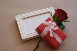 Top view of red rose and red gift box laying on a white blank photo frame isolated on brown background. Valentine's day, wedding, birthday and special occasion concept. Flat lay, top view.