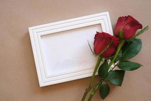 Top view of red rose and red heart laying on a white blank photo frame isolated on brown background. Valentine's day, wedding, birthday and special occasion concept. Flat lay, top view.